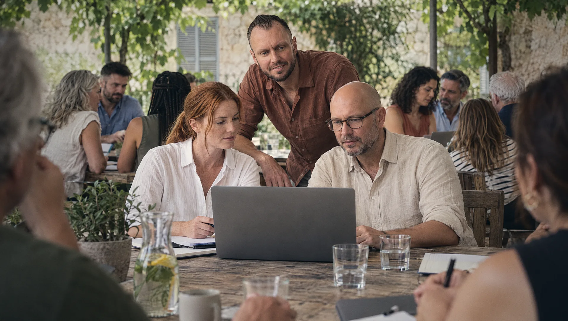 Michael Selck arbeitet mit zwei Unternehmern am Laptop, an den Nachbartischen unter der Pergola arbeiten weitere Gruppen — Bestandsaufnahme in der Digitalisierungsberatung